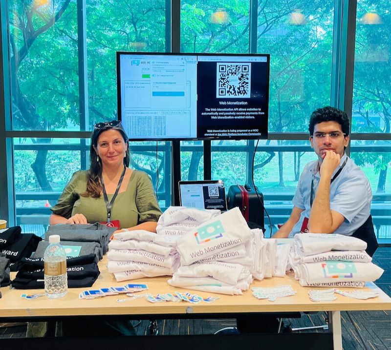 Ioana and Sid at the booth, with demo in background and lots of tshirts in front. Photo by @devcer